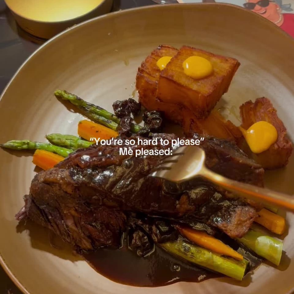 Plated roasted beef with asparagus, carrots, glazed vegetables, crispy potato cubes, and yellow sauce in a warmly lit restaurant setting.