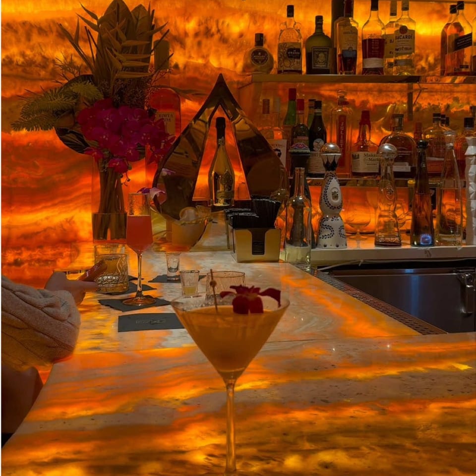 Orange-lit cocktail bar with liquor bottles, tropical flowers, and a martini garnished with a cherry on the marble counter.
