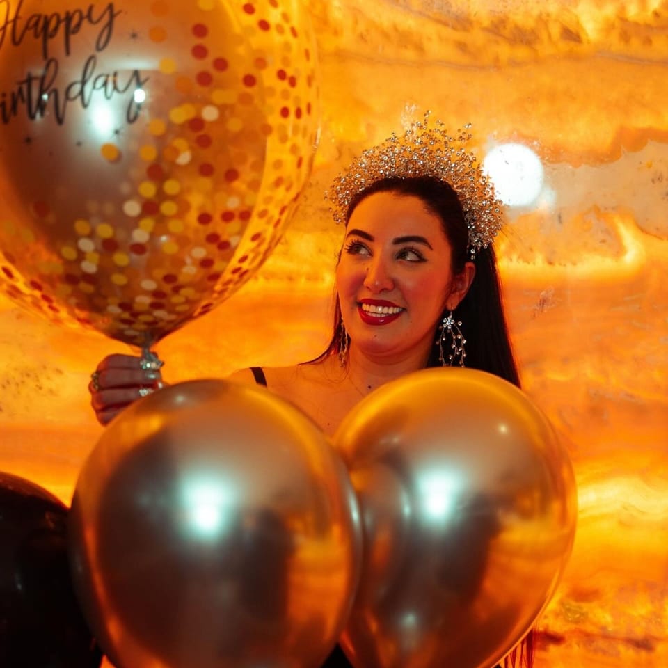Smiling woman wearing a sparkling tiara poses among orange and gold balloons against a warm, festive backdrop.