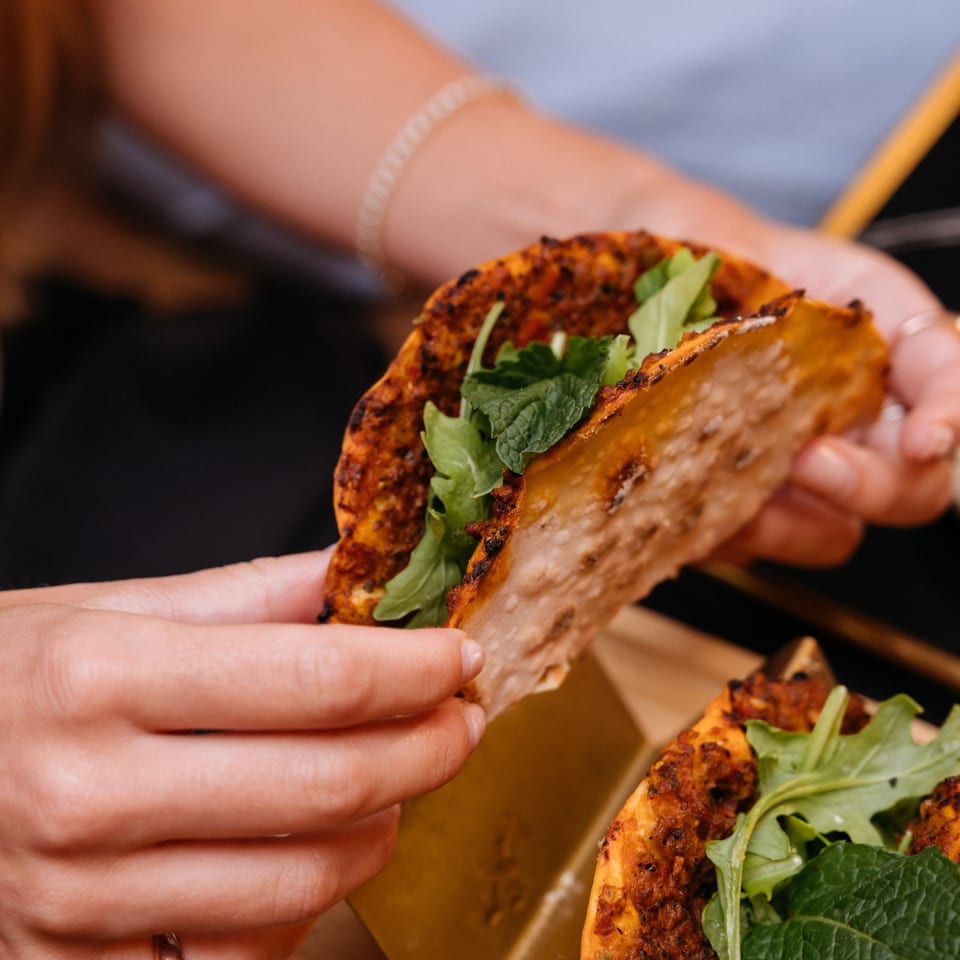 Hands hold a crispy folded taco filled with grilled meat, greens, and herbs above a tray of additional tacos.