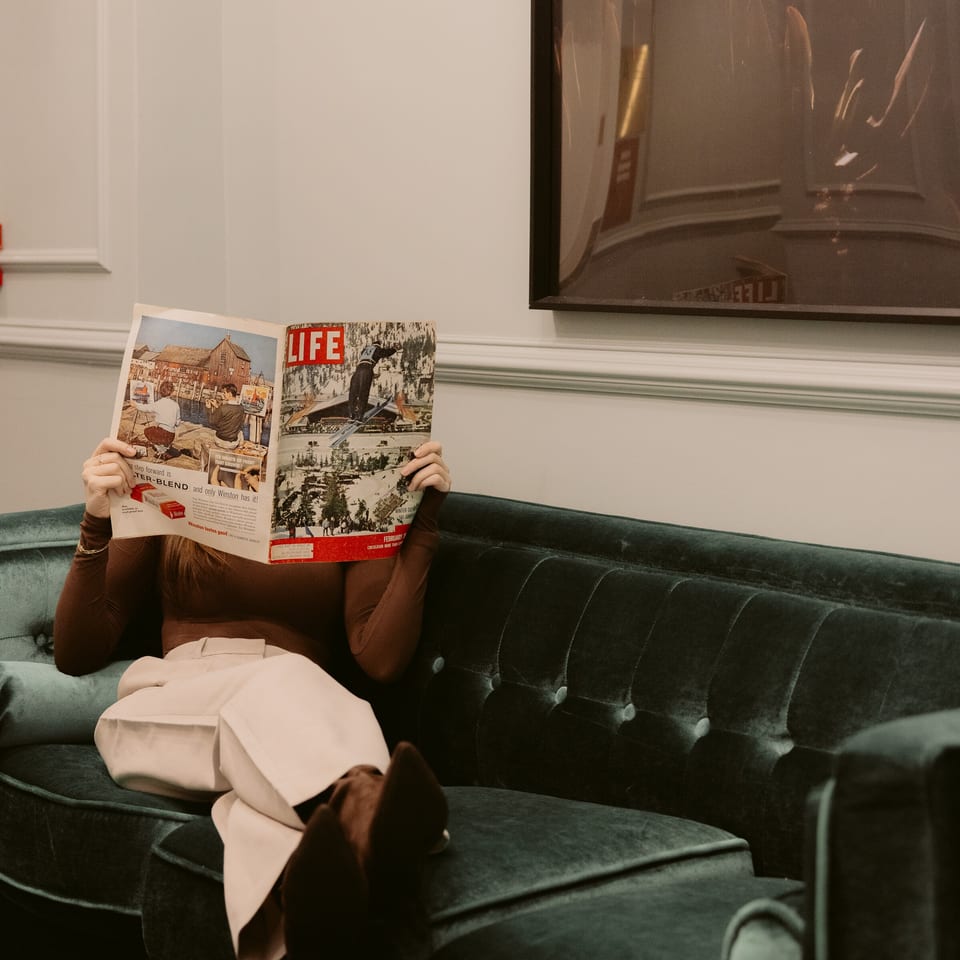 Person reclining on a dark green velvet sofa, reading a magazine in an elegant room with framed artwork.