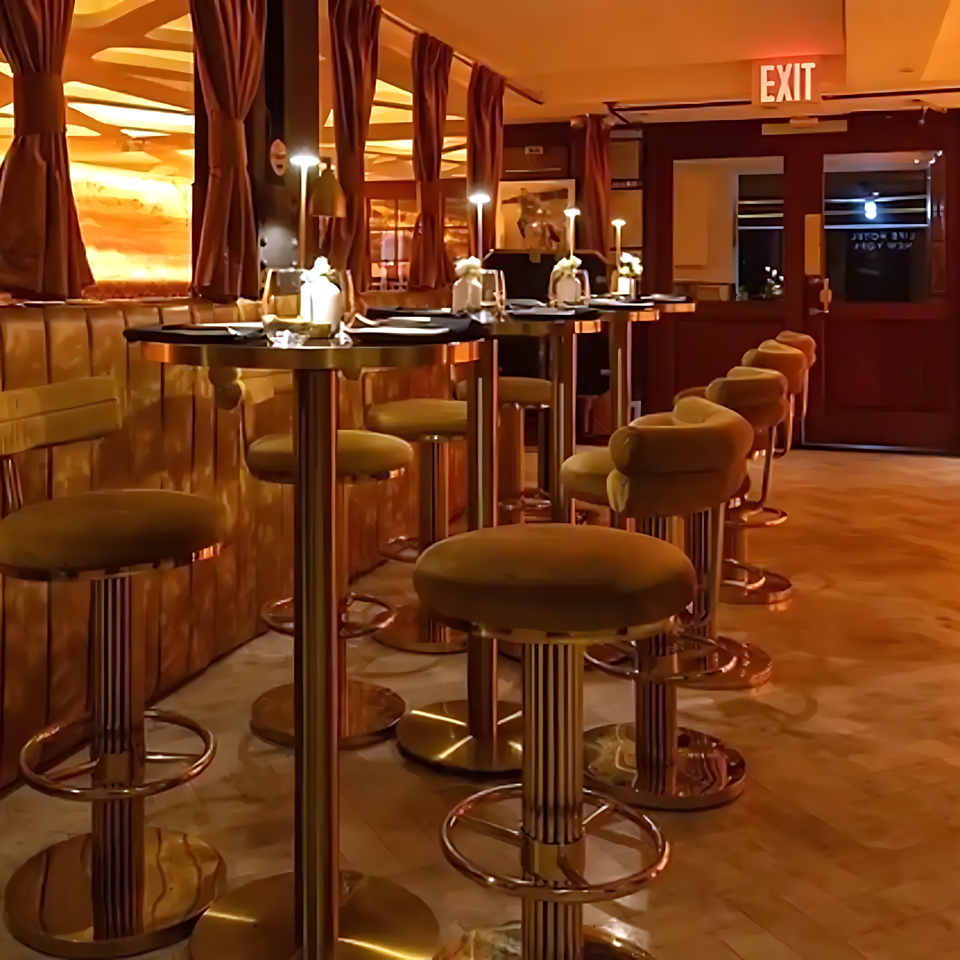 Warmly lit restaurant lounge with upholstered booths, marble-topped cocktail tables, bar stools, draped curtains, and an exit door.