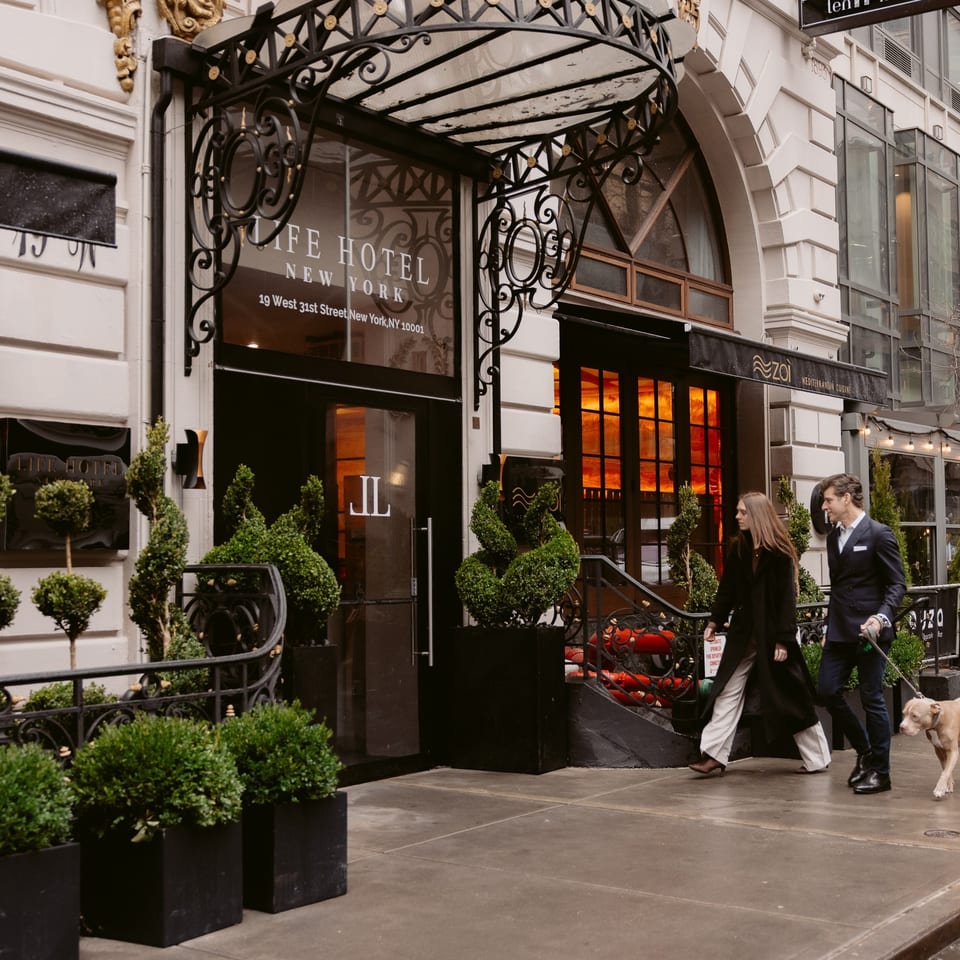 Cafe Hotel New York entrance with ornate iron canopy, manicured topiaries, outdoor dining, and two people walking a dog.