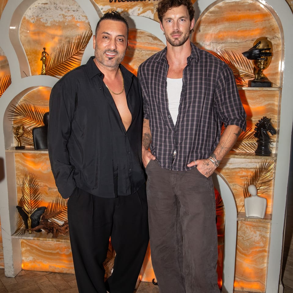 Two men pose inside ZOI Mediterranean restaurant, surrounded by orange stone walls, tropical decor, and sculptural artwork.