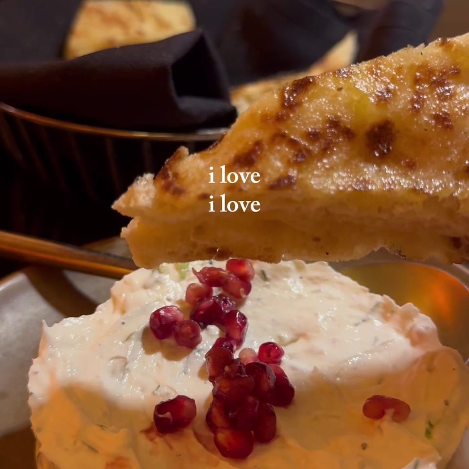 Crispy toasted flatbread rests over creamy dip garnished with pomegranate seeds, with bread basket and cutlery in the background.