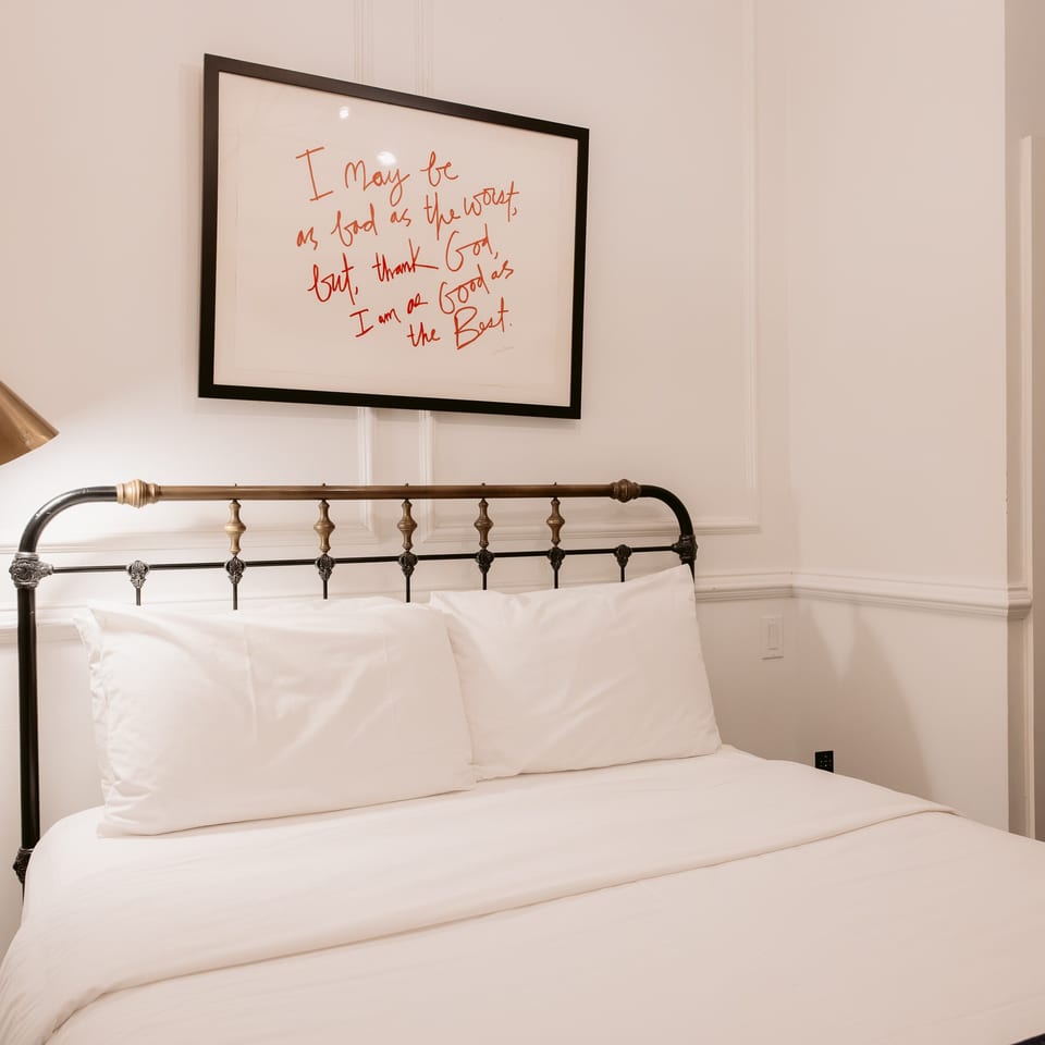 Bright bedroom with a neatly made white bed, black metal headboard, wall-mounted reading lamp, framed handwritten artwork, and paneled walls.