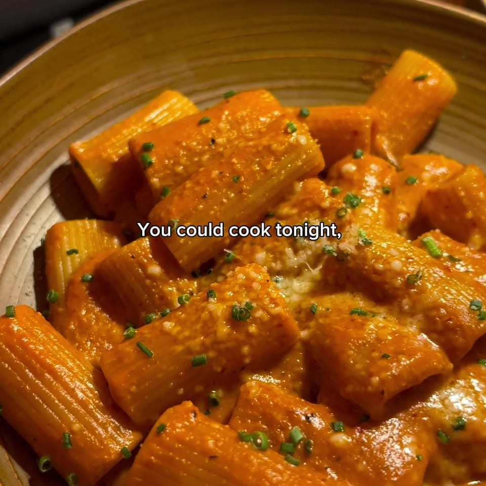 Bowl of rigatoni coated in creamy orange sauce and sprinkled with chopped green herbs, beside a fork, knife, and water glass.