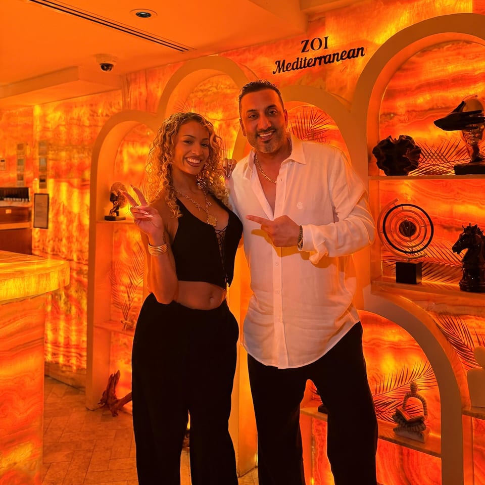 Two smiling people pose inside ZOI Mediterranean, surrounded by warm orange lighting, arched decor, and sculptural artwork.