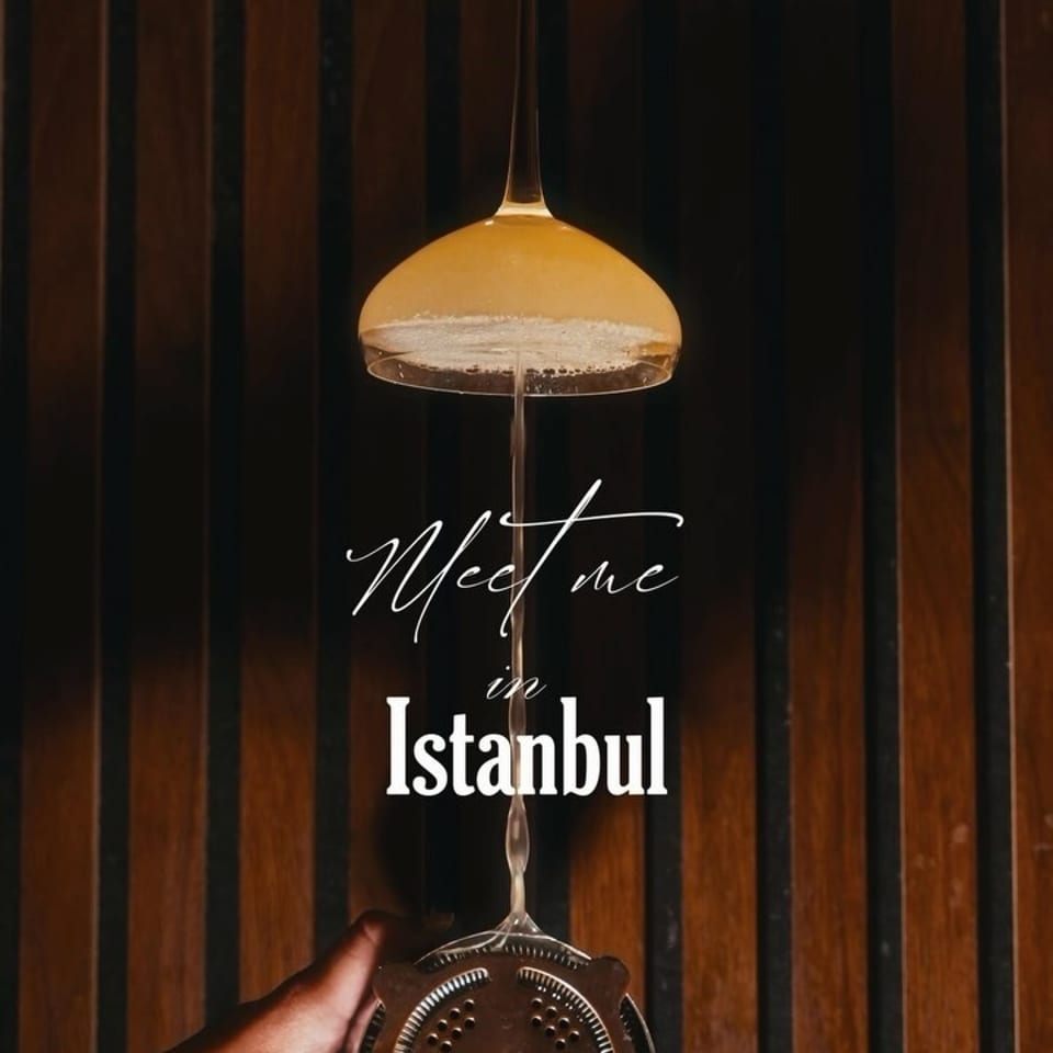 Hands strain a golden cocktail through a coupe glass against dark wood, with text reading “Meet me in Istanbul.”