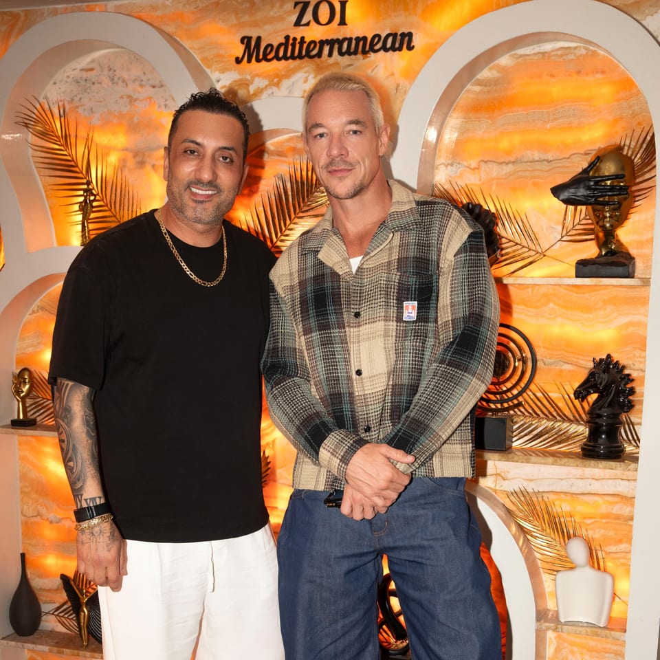 Two men pose beneath the ZOI Mediterranean sign in a warmly lit restaurant featuring orange stone walls, palm decor, and sculptures.