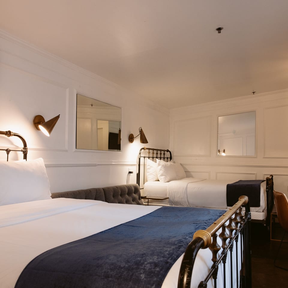 Bright boutique hotel room with two brass-framed beds, navy runners, tufted seating, wall mirrors, and a desk.