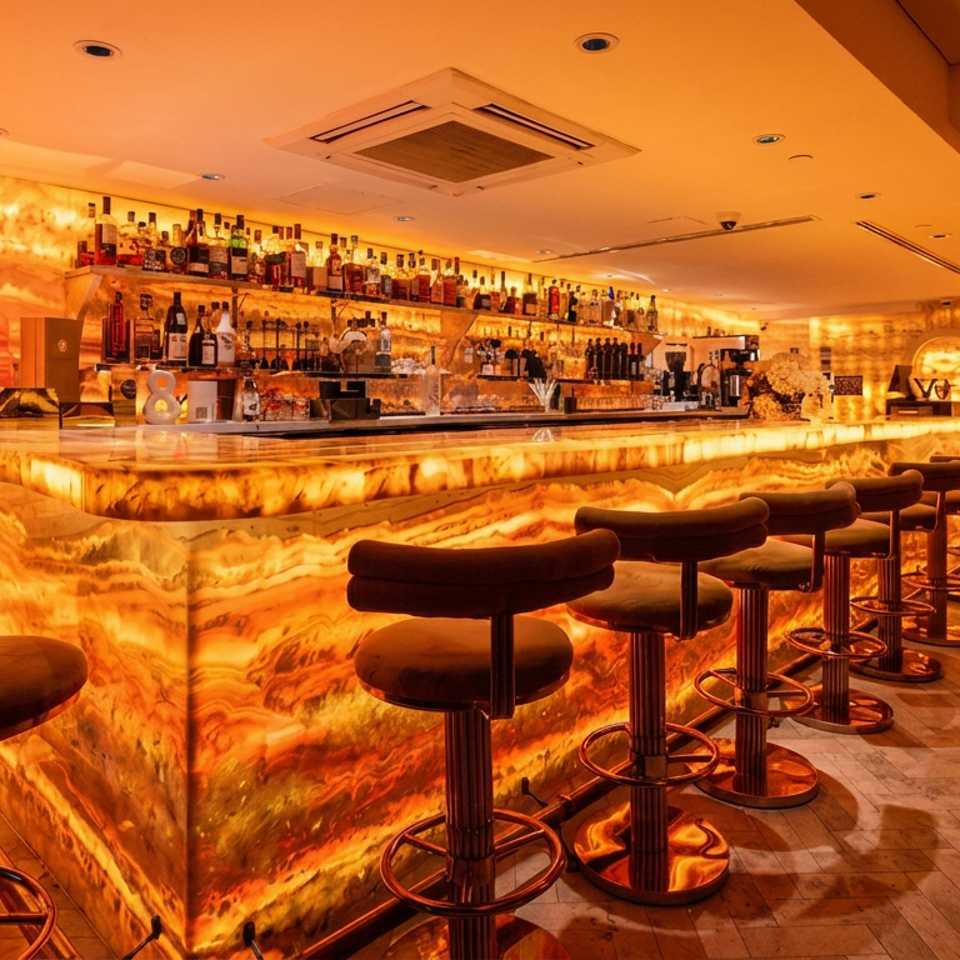 Warmly lit upscale cocktail bar with glowing marble counters, liquor shelves, sculptural wall niches, and rows of velvet bar stools.