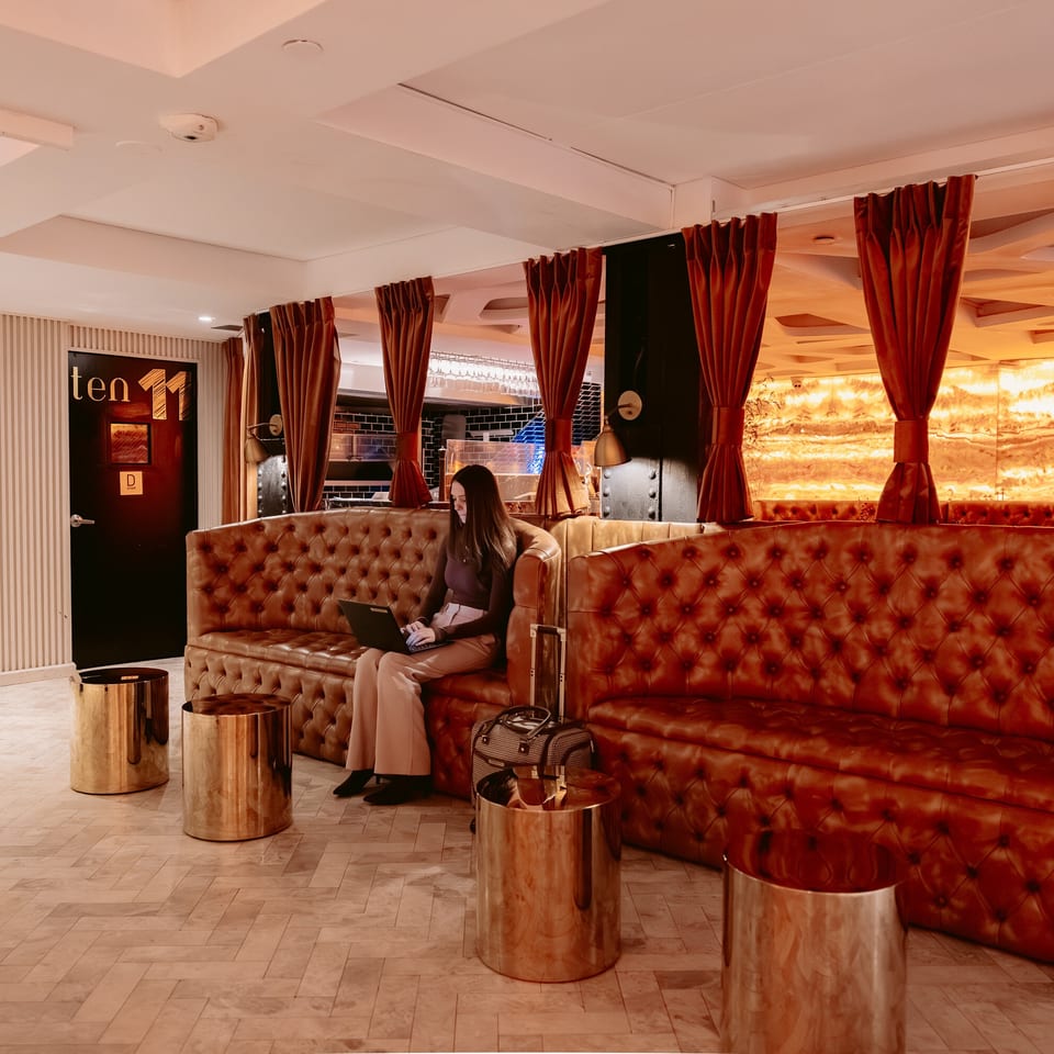 Warmly lit restaurant lounge with tufted leather sofas, red curtains, brass tables, and a woman using a laptop.