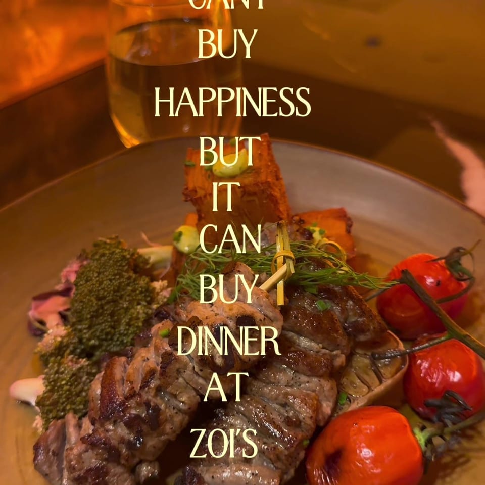 Grilled meat skewers with roasted tomatoes and vegetables on a plate at Zoi’s, with a cocktail and menu in the background.