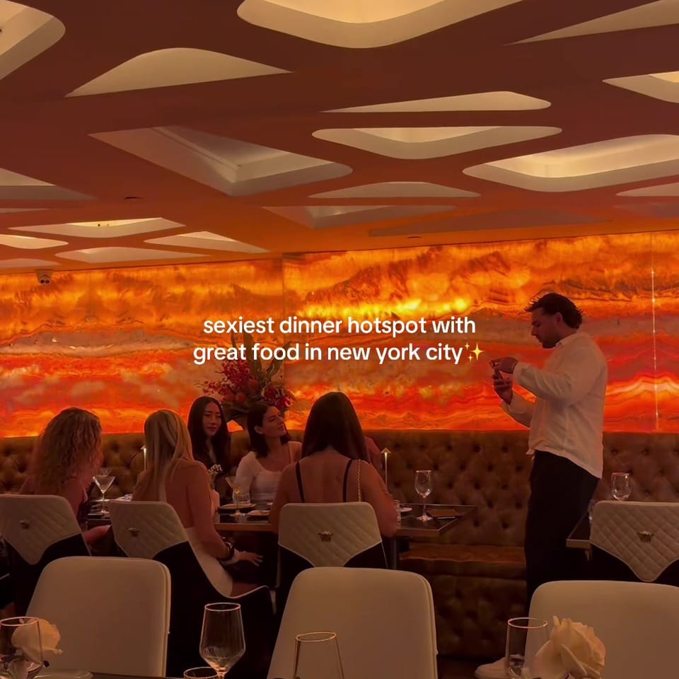 Upscale restaurant dining room with orange illuminated walls, sculptural ceiling lights, seated diners, and a standing server.