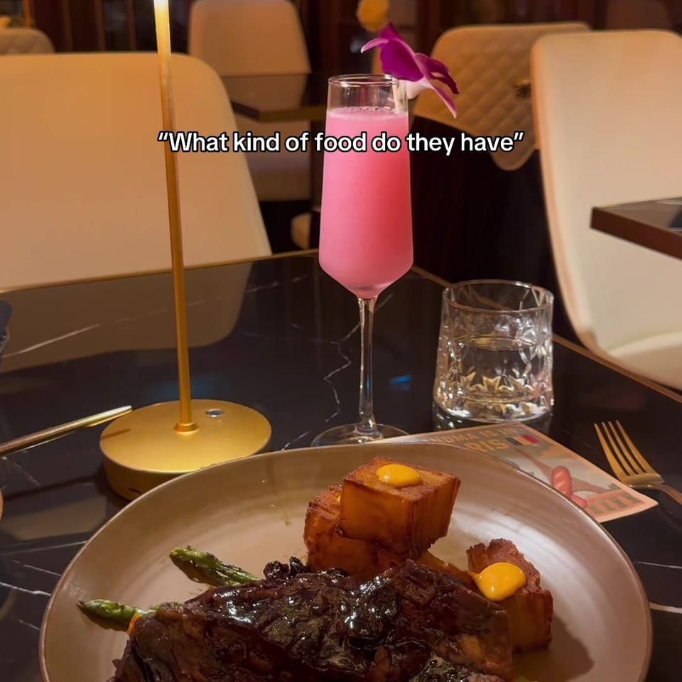 Upscale restaurant table with glazed steak, roasted vegetables, pink cocktail, water glass, and a small table lamp.