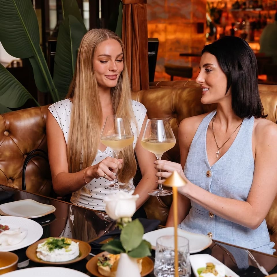 Two women toast with white wine in a warmly lit restaurant booth surrounded by plated dishes and floral décor.