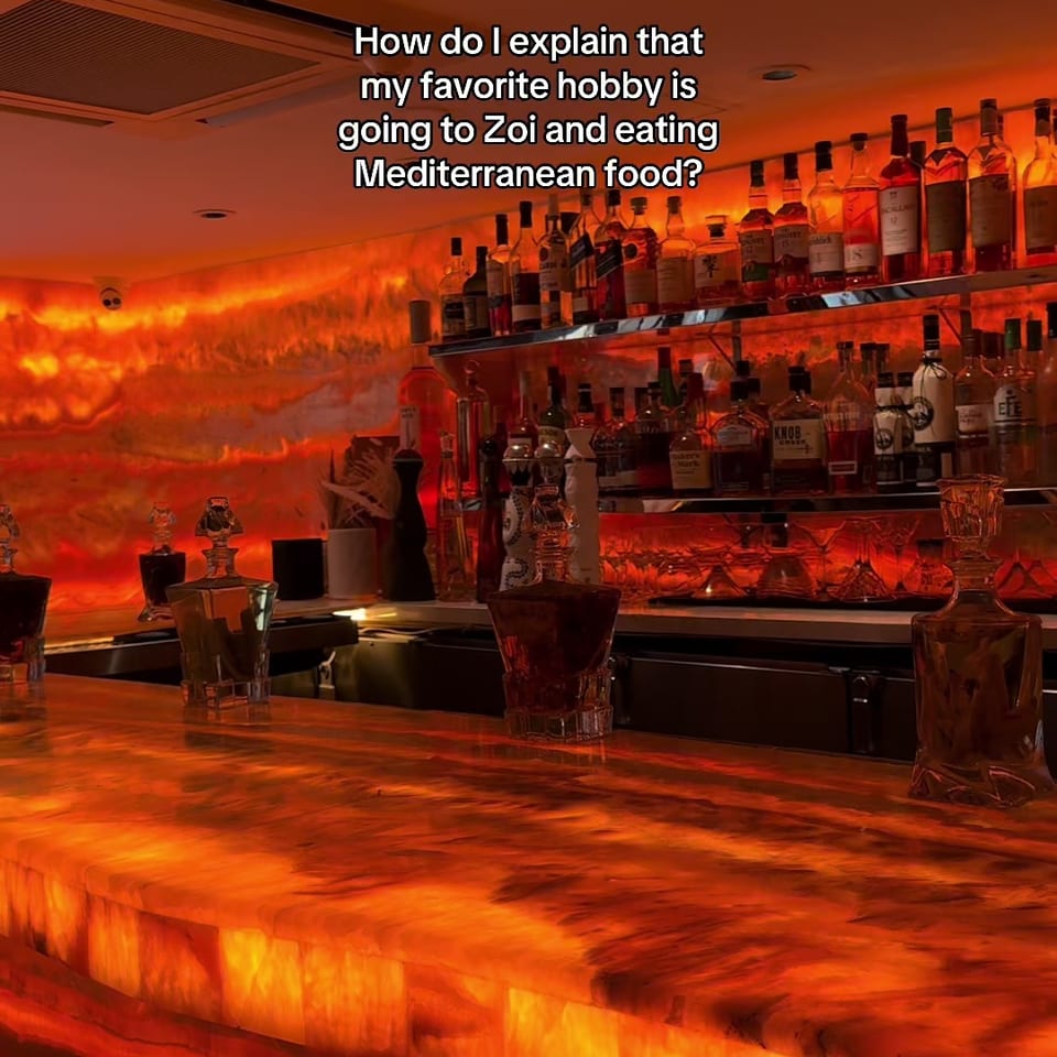 Warmly lit Zoi restaurant bar with liquor shelves, glowing orange stone counters, and Mediterranean dining atmosphere.