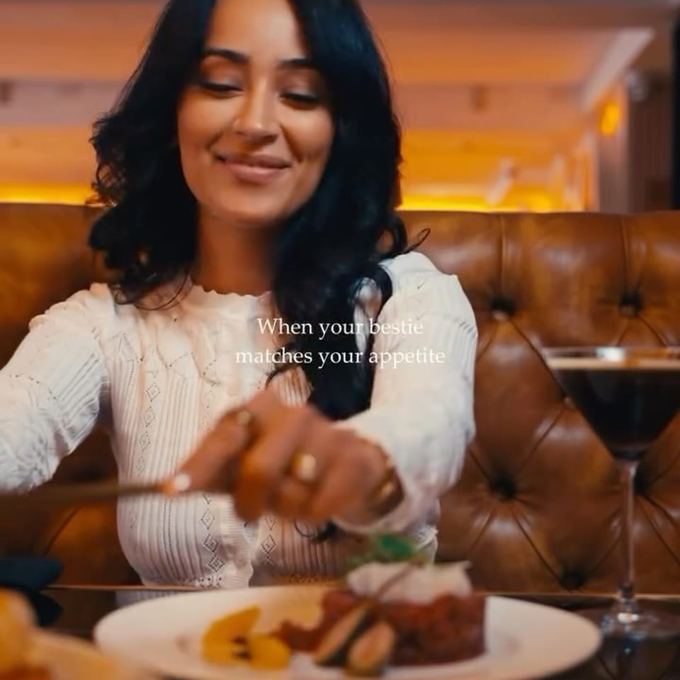 Woman dining in a warmly lit restaurant booth, reaching toward a plated gourmet meal beside a glass of red wine.